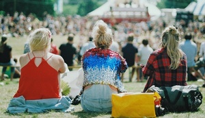Menschen sitzen bei Sonnenschein auf einer Wiese und blicken auf ein Zelt bzw. eine Bühne.