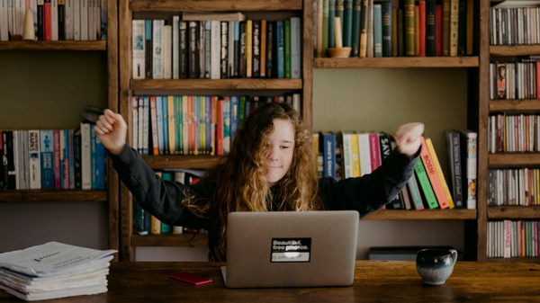 Mädchen vor Laptop, freudig entspannt die Arme streckend. Bücherwand im Hintergrund, © Annie Spratt@unsplash Mädchen vor Laptop, freudig entspannt die Arme streckend. Bücherwand im Hintergrund, © Annie Spratt@unsplash