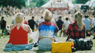 Menschen sitzen bei Sonnenschein auf einer Wiese und blicken auf ein Zelt bzw. eine Bühne.