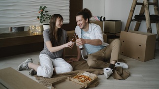 am Boden sitzendes junges Paar mit Pizza und Getränken, umgeben von Umzugskisten 
